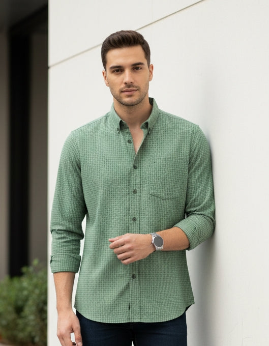 Man wearing a green shirt and dark jeans standing against a white wall.